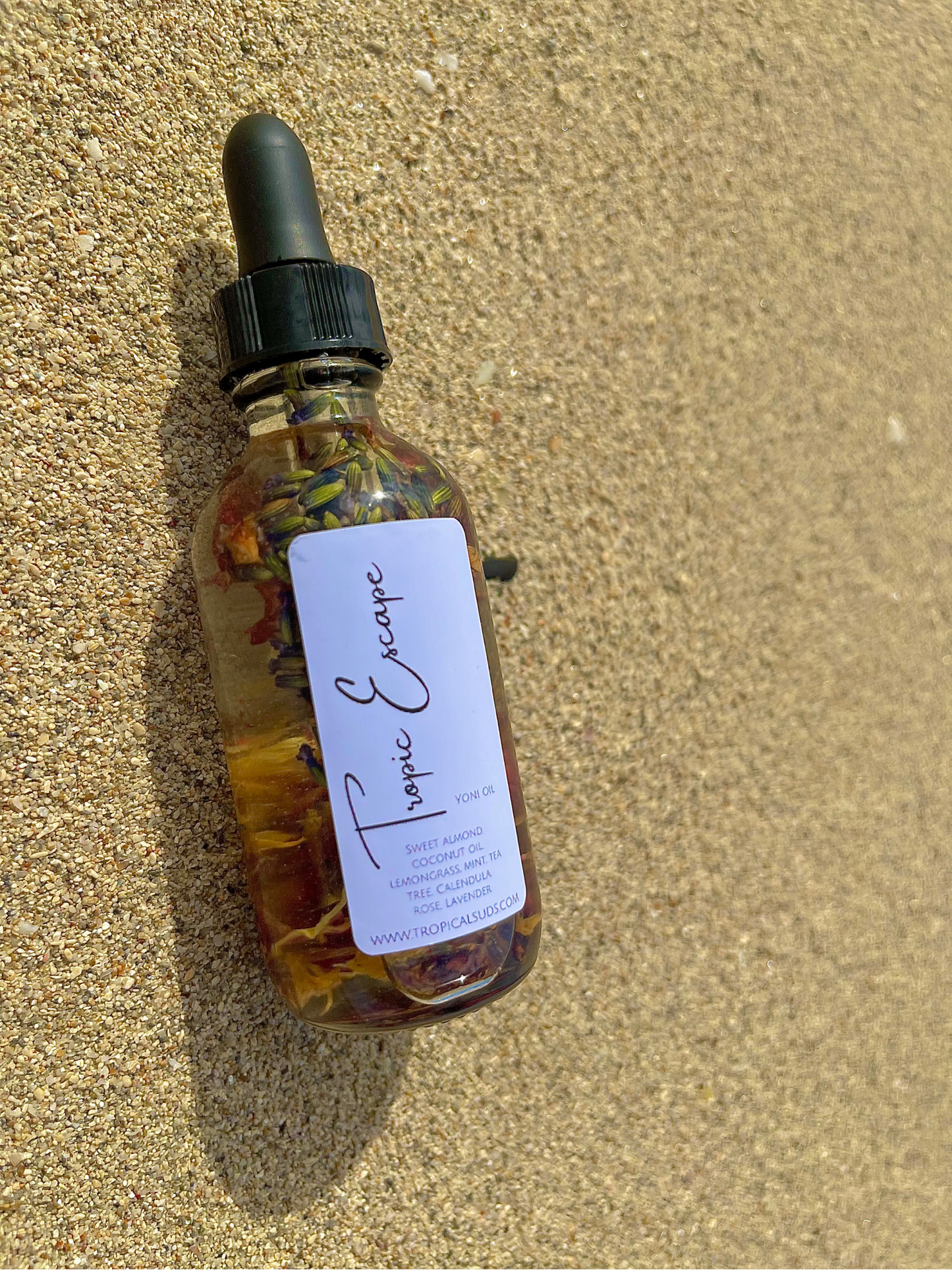 A clear glass bottle with a dropper filled with yellowish liquid, filled with the Tropic Escape Oil, placed on a sandy surface.